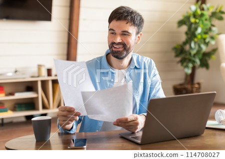 A man is seated at a table, holding a piece of paper in his hands. He appears focused on the paper, potentially reading or writing. 114708027