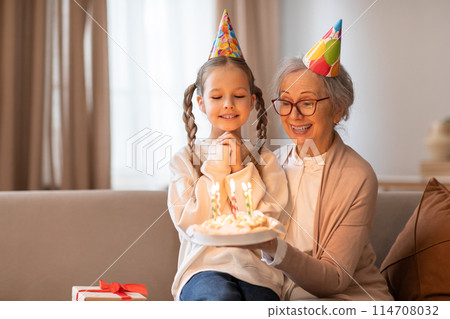 An older woman and a young girl sit on a couch, with a birthday cake in front of them. The woman is lighting the candles on the cake while the girl eagerly watches An older woman and a young girl sit on a couch, with a birthday cake in front of them. The woman is lighting the candles on the cake while the girl eagerly watches 114708032