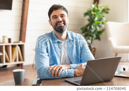 A man is sitting at a desk, facing a laptop computer with a serious expression. He is typing on the keyboard, seemingly focused on his work or task at hand. 114708040