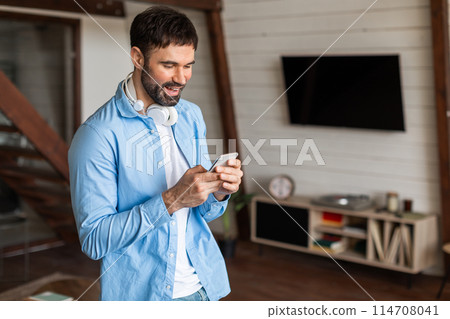 A man is standing in a living room, focused on his cell phone screen. He appears engaged in whatever he is observing or reading on the device. A man is standing in a living room, focused on his cell phone screen. He appears engaged in whatever he is observing or reading on the device. 114708041