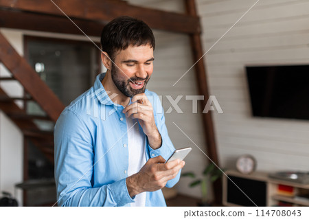 A cheerful young man with a beard is seen holding and looking at his smartphone with a smile, suggesting he has received good news 114708043