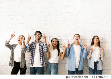 A diverse group of five multiethnic teenagers is standing side by side against a white brick wall, all smiling and looking upwards while simultaneously pointing with their index fingers A diverse group of five multiethnic teenagers is standing side by side against a white brick wall, all smiling and looking upwards while simultaneously pointing with their index fingers 114708052