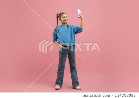 A cheerful young girl stands confidently with one hand on her hip and the other holding a smartphone above her head, capturing a selfie A cheerful young girl stands confidently with one hand on her hip and the other holding a smartphone above her head, capturing a selfie 114708092