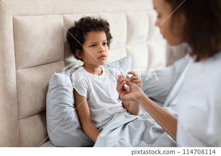 African American young child sits with a contemplative expression on bed while an adult, partially visible, holds out a pill for him to take, likely indicating a moment of healthcare at home. African American young child sits with a contemplative expression on bed while an adult, partially visible, holds out a pill for him to take, likely indicating a moment of healthcare at home. 114708114