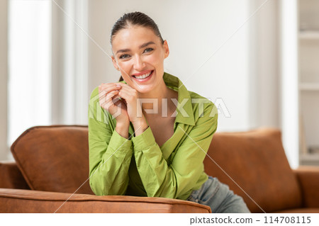 A woman is seated on a couch, smiling directly at the camera. She appears relaxed and content in her expression, exuding warmth and approachability. 114708115
