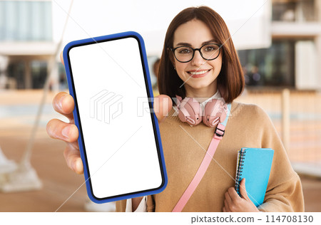 A woman student is holding up a modern smart phone with a blank screen in her hand. She appears to be looking at the screen, potentially preparing to use it for communication or information. A woman student is holding up a modern smart phone with a blank screen in her hand. She appears to be looking at the screen, potentially preparing to use it for communication or information. 114708130
