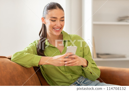 A woman is seated on a couch, engrossed in her cell phone. Her attention is solely focused on the device in her hands. A woman is seated on a couch, engrossed in her cell phone. Her attention is solely focused on the device in her hands. 114708132