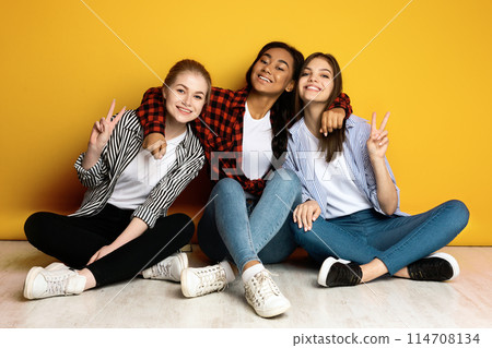 Three young girls are sitting on the floor, striking different poses as if they are getting ready for a group photo. They appear cheerful and excited, forming various poses with their arms and legs. 114708134