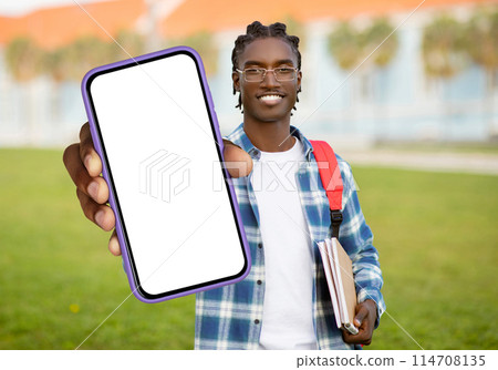 A cheerful young black man with braided hair carrying a red backpack, holds out a smartphone with a blank screen while standing on a university campus with buildings in the background A cheerful young black man with braided hair carrying a red backpack, holds out a smartphone with a blank screen while standing on a university campus with buildings in the background 114708135