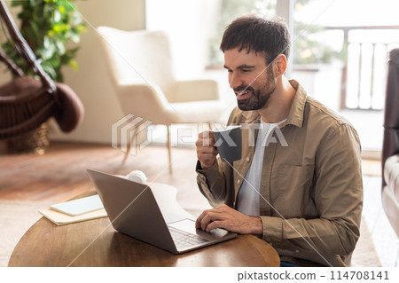 A man in a casual attire is sitting at a table, focusing on his laptop screen. Beside him, there is a cup of coffee, suggesting a working environment 114708141