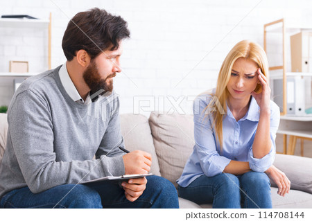A seated man, possibly a counselor or therapist, is attentively listening to a woman who appears distressed or concerned. A seated man, possibly a counselor or therapist, is attentively listening to a woman who appears distressed or concerned. 114708144