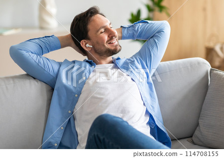 A content man reclines comfortably on a gray sofa with his hands behind his head, eyes closed, and a pleased smile, as he listens to music through his white wireless earbuds. 114708155