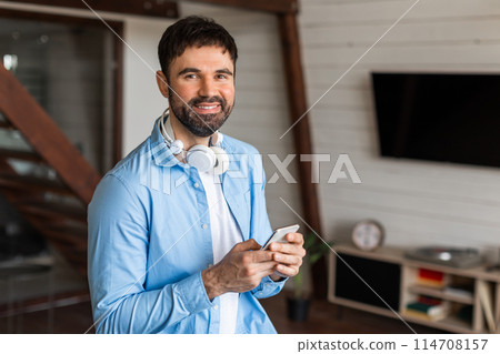 A man is captured wearing headphones, listening to music, standing in a casual blue shirt. He appears to be engaged in his music and relaxed. 114708157