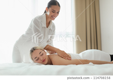 A woman is lying face down on a massage table while another woman, dressed in a white shirt, is giving her a back massage. The setting appears to be a professional massage studio. 114708158
