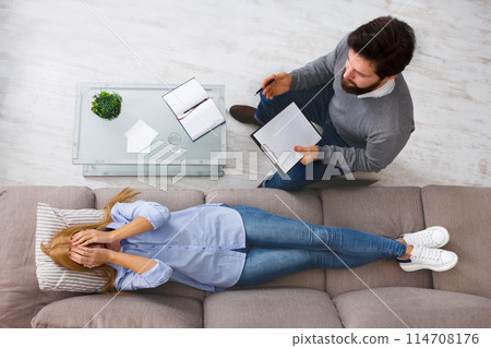 A bearded man therapist is attentively listening to woman client as she lies on a couch, sharing her thoughts during a psychotherapy session, view above 114708176