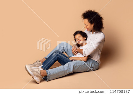 African American young mother sits on the floor, cradling her child in her arms with a warm embrace. The background is a plain, soft beige 114708187