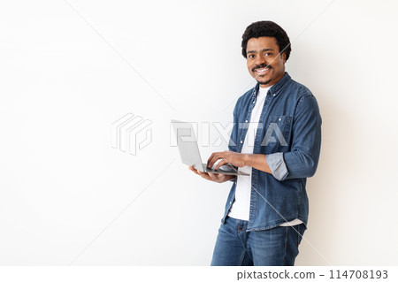 African American man is standing, holding a laptop in his hands. He is looking at the screen intently, possibly working or browsing. The background is white with copy space 114708193