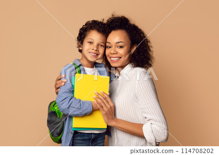 A cheerful African American mother lovingly hugs her young son, who is prepared for a day at school with his backpack and a bright yellow notebook 114708202