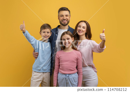 A cheerful family, with two children and their parents, stand close together with bright smiles, each raising their thumb in a sign of approval or success 114708212