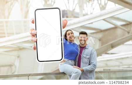 A man and woman are standing together, holding up a cell phone with white blank screen, mockup copy space. Traveling app A man and woman are standing together, holding up a cell phone with white blank screen, mockup copy space. Traveling app 114708234