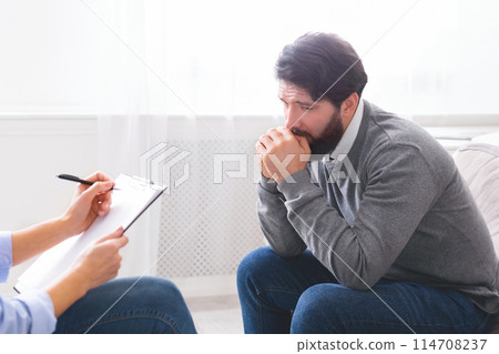 A distressed man is seen with his hand on his mouth as if in deep thought or concern, talking to a counselor who is taking notes. They are seated across from each other in a well-lit office space A distressed man is seen with his hand on his mouth as if in deep thought or concern, talking to a counselor who is taking notes. They are seated across from each other in a well-lit office space 114708237