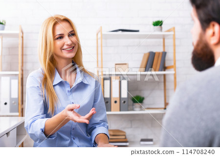 In an office setting, a woman is engaged in conversation with a man. They appear to be discussing business matters, with papers and a computer visible on the desk in front of them. 114708240