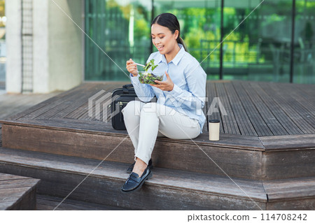 An Asian businesswoman is sitting on steps outdoors while eating a salad. She is dressed professionally and appears to be taking a break from work to enjoy her meal. 114708242
