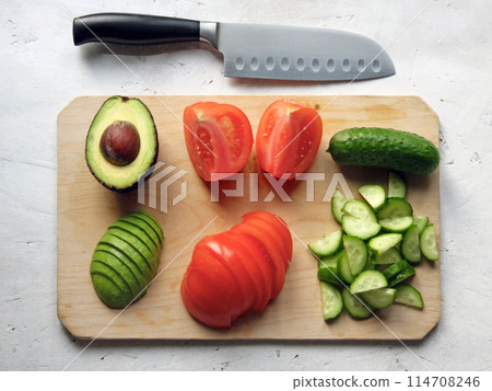 An avocado with a stone, a tomato and a cucumber cut into slices lie on a white table on a wooden board next to a knife in the kitchen. Fresh clean vegetables, farming, salad preparation, rustic An avocado with a stone, a tomato and a cucumber cut into slices lie on a white table on a wooden board next to a knife in the kitchen. Fresh clean vegetables, farming, salad preparation, rustic 114708246