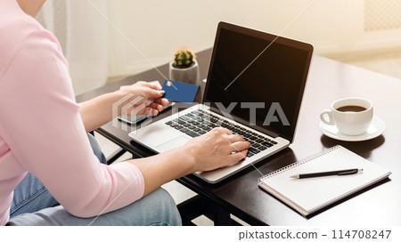 A woman sits at a cafe table engaging in online shopping or payment processing, while holding her credit card in one hand and typing on her laptop with the other. 114708247