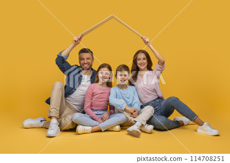 A happy family composed of a father, a mother, a daughter, and a son is sitting together on a yellow background, smiling as they hold a white picture frame above their heads A happy family composed of a father, a mother, a daughter, and a son is sitting together on a yellow background, smiling as they hold a white picture frame above their heads 114708251