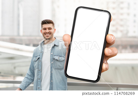 A man is holding a smart phone with white blank screen in his hand. The background is neutral, focusing on the man and the technology in his hand. 114708269