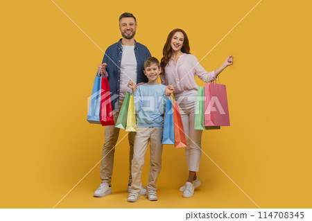 A cheerful family of three, comprising a father, mother, and their young son, stands confidently against a bright yellow background, each holding several colorful shopping bags 114708345