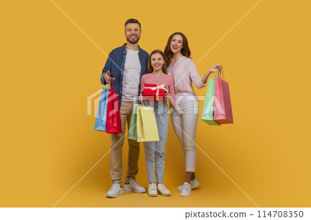 A happy family consisting of a father, mother, and their young daughter pose together with bright shopping bags and gift in their hands A happy family consisting of a father, mother, and their young daughter pose together with bright shopping bags and gift in their hands 114708350