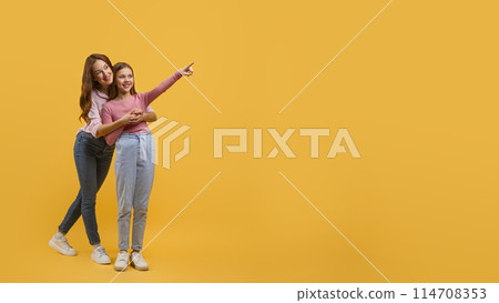 Mother and daughter dressed in casual wear, stand closely together against a vibrant yellow backdrop, pointing at copy space while they both smile brightly, sharing a lighthearted moment. Mother and daughter dressed in casual wear, stand closely together against a vibrant yellow backdrop, pointing at copy space while they both smile brightly, sharing a lighthearted moment. 114708353