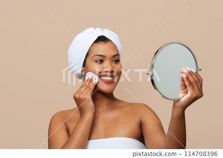 A cheerful young woman wrapped in a white towel looks at her reflection in a hand-held mirror while applying tonner to her cheek. Her hair is tucked under a white headband 114708396