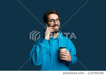 A cheerful young man with a beard and glasses, dressed in a casual blue hoodie, engages in a conversation on his mobile phone while simultaneously holding a takeaway coffee cup A cheerful young man with a beard and glasses, dressed in a casual blue hoodie, engages in a conversation on his mobile phone while simultaneously holding a takeaway coffee cup 114708398