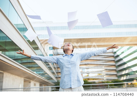 Chinese woman is energetically tossing papers into the air outside an office building. The scene is filled with flying papers against the backdrop of a corporate skyscraper. Chinese woman is energetically tossing papers into the air outside an office building. The scene is filled with flying papers against the backdrop of a corporate skyscraper. 114708440