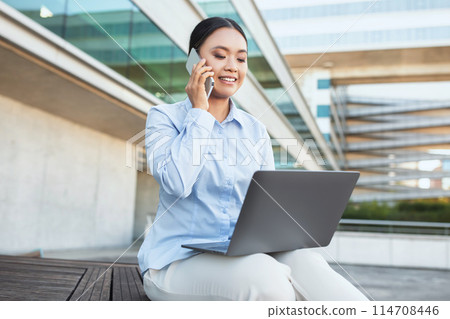 Asian woman is seated on a bench, engrossed in a phone conversation while simultaneously working on a laptop. She appears focused and multitasking efficiently in a public setting. 114708446
