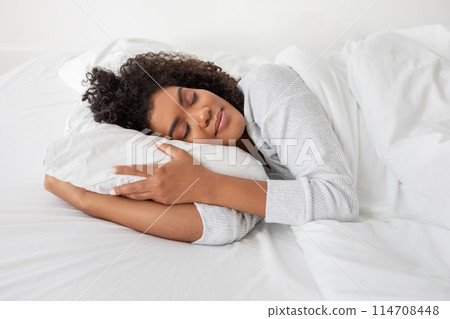 Hispanic woman is seen laying in bed, with a pillow under her head. She appears comfortable and relaxed, possibly resting or sleeping, side view 114708448