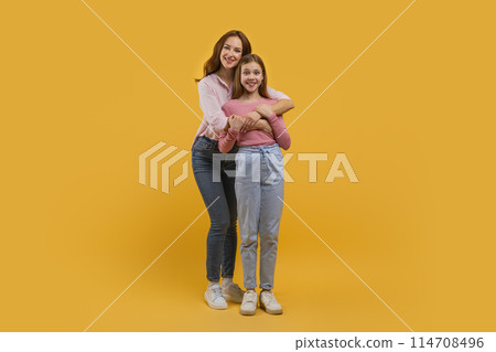 Mother and daughter stand closely with one embracing the other from behind. Both are smiling warmly and dressed casually in jeans and light tops against a vivid yellow backdrop. 114708496
