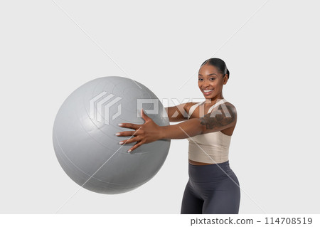 African American woman is seen holding a huge exercise ball in a gym. She is wearing workout attire, and her posture suggests she is preparing for a fitness routine African American woman is seen holding a huge exercise ball in a gym. She is wearing workout attire, and her posture suggests she is preparing for a fitness routine 114708519