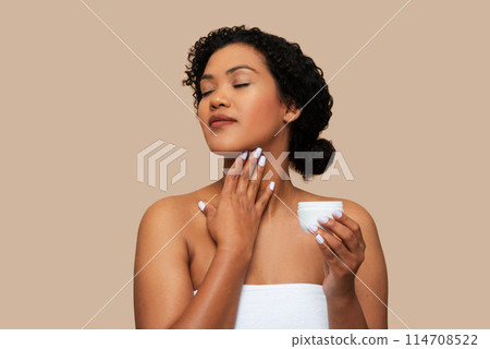 A young woman with curly hair, wrapped in a white towel, gently applies moisturizing cream to her neck, moment of self-care and relaxation. The neutral beige background 114708522