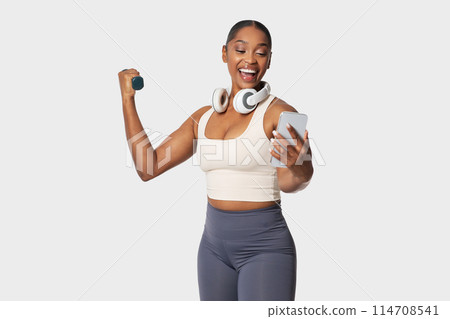 A cheerful young African American woman in athletic wear is holding a dumbbell in one hand and a smartphone in the other, seemingly following a workout routine 114708541
