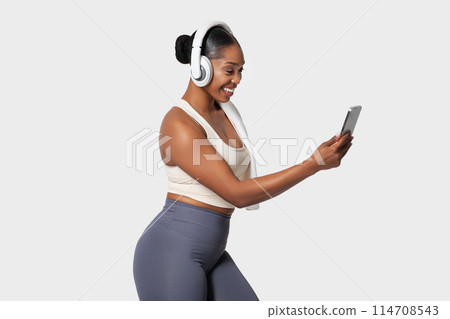 A cheerful African American woman is engaged with phone, wearing a pair of over-ear headphones, dressed in casual workout attire. Her attention is focused on the screen A cheerful African American woman is engaged with phone, wearing a pair of over-ear headphones, dressed in casual workout attire. Her attention is focused on the screen 114708543