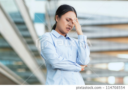 Asian woman in business attire appears distressed and holds her head, signaling a headache or overwhelming stress, against the backdrop of a modern urban environment Asian woman in business attire appears distressed and holds her head, signaling a headache or overwhelming stress, against the backdrop of a modern urban environment 114708554