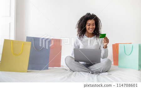 A cheerful young Hispanic woman is seated on bed with her legs crossed, surrounded by colorful shopping bags, holding a credit card in hand while looking at her laptop screen 114708601