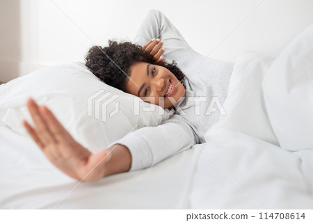 A cheerful Hispanic woman is lying in bed, with her head resting on a pillow and a comfortable smile on her face. She is casually dressed in cozy, light sleepwear, reaching out towards the camera 114708614