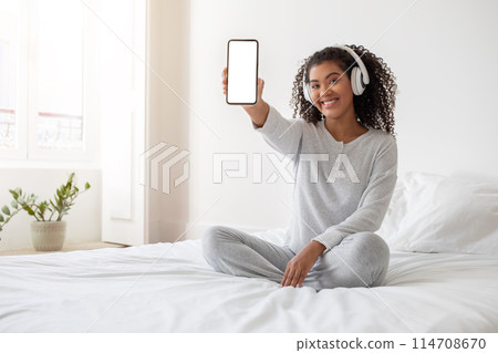 A cheerful Hispanic woman sits cross-legged on a white bed, wearing casual grey clothing and headphones. She is holding up her smartphone facing forward, displaying a blank screen A cheerful Hispanic woman sits cross-legged on a white bed, wearing casual grey clothing and headphones. She is holding up her smartphone facing forward, displaying a blank screen 114708670
