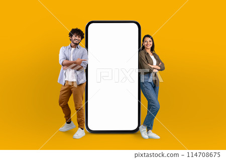 A cheerful young hindu man and woman stand confidently next to a large, blank screen smartphone that dominates the center of the frame, suggesting a technology theme 114708675