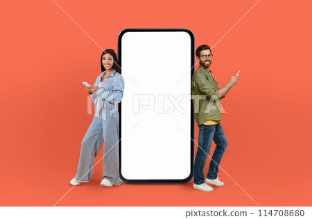 Multiracial lovers standing side by side next to a landline phone. They both appear to be looking at something on the phone screen, with expressions of concentration and interest. 114708680